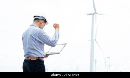 Il successo ingegnere mulini a vento indossando maschera e lavorando sul computer portatile con la turbina eolica in background Foto Stock