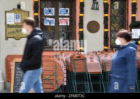 Monaco, Germania. 05 maggio 2021. Soggetto: Pandemia di coronavirus/conseguenze per la gastronomia: RISTORANTE APERTO IMMEDIATAMENTE nella finestra di un ristorante bavarese a Monaco, i proprietari chiedono l'apertura immediata della gastronomia. Sedie e tavoli impilati uno sull'altro si appoggiano contro il muro della casa. Passers-by pass by con maschera facciale, maschera. | utilizzo in tutto il mondo Credit: dpa/Alamy Live News Credit: dpa Picture Alliance/Alamy Live News Foto Stock