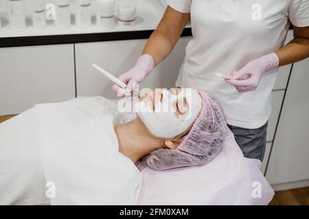 Cosmetologist che applica la maschera sul viso del cliente nel salone termale. Giovane donna che ottiene la cura del viso da estetista al salone termale trattamento Acne, peeling del viso Foto Stock