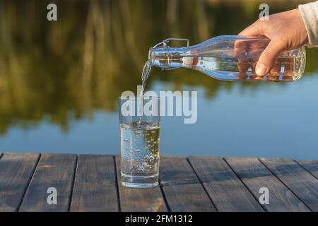Versare l'acqua in un bicchiere da una bottiglia la mattina accanto al lago e alla foresta in primavera, primo piano. Natura e concetto di viaggio Foto Stock