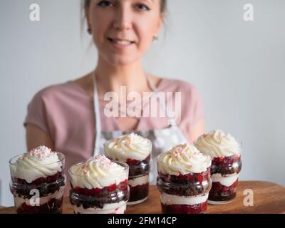 Happy cuoco femmina nel grembiule tiene un piatto con trifles di ciliegia fresco, fondo grigio. Foto Stock
