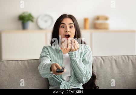 Scioccata donna indiana seduta sul divano, mangiando popcorn e guardando un eccitante programma TV o film a casa Foto Stock