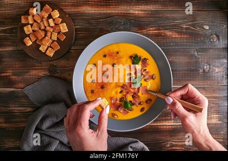 Le mani della donna tengono il crostino e il cucchiaio con la tradizionale zuppa di crema purea di zucca con carote, cipolle, zenzero, aglio, latte di cocco. Aggiunta di semi, pancetta fritta, prezzemolo. Cibo sano e vegetariano. Vista dall'alto Foto Stock