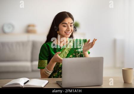 Donna indiana positiva in abito sari tradizionale indossando cuffie, avendo riunione on-line sul computer portatile a casa Foto Stock