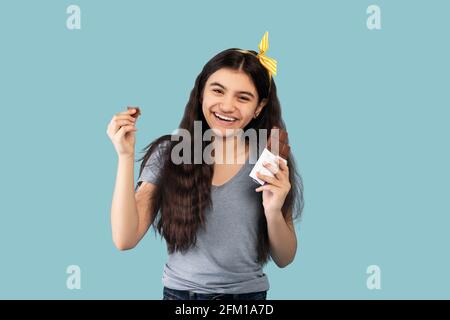 Ritratto di felice ragazza indiana teen mangiare cioccolato bar, sensazione di piacere su sfondo blu studio Foto Stock