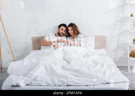coppia interrazziale sorridente mentre si sdraiava a letto e si usa uno smartphone Foto Stock