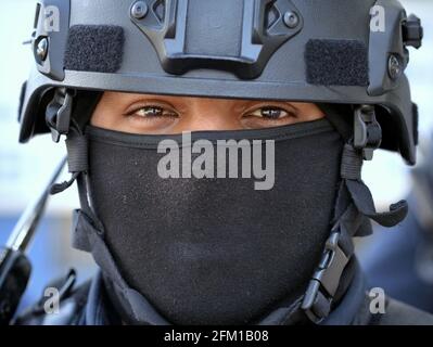 Il giovane ufficiale di polizia del motociclo tattico caucasico indossa i dispositivi di protezione personali neri con il casco e la maschera nera del viso e guarda lo spettatore. Foto Stock