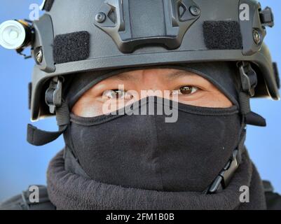 Il giovane e duro ufficiale di polizia del motociclo tattico indossa i dispositivi di protezione personali e una balaclava nera e guarda con gli occhi stanchi allo spettatore. Foto Stock