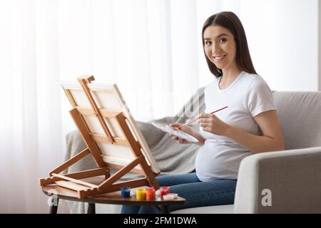 Passatempo durante la gravidanza. Bella donna incinta pittura su Easel a casa Foto Stock