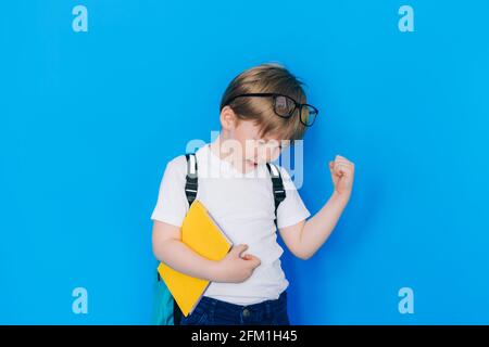 Concetto di ritorno a scuola. Scolaro con zaino e taccuino giallo contro la parete blu. Bambino pugno clenched che mostra successo. Foto Stock