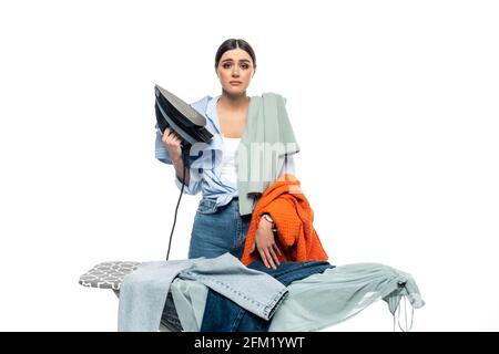 donna sconvolsa che tiene il ferro rotto vicino all'asse da stiro e ai vestiti isolato su bianco Foto Stock