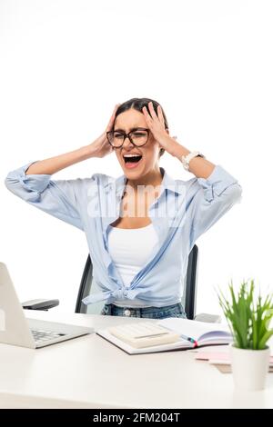 irritata donna d'affari che grida e tocca la testa mentre si siede vicino al computer portatile isolato su bianco Foto Stock