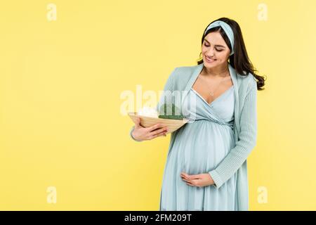 gioiosa donna incinta che tiene la ciotola con cavolfiore fresco e broccoli isolato in giallo Foto Stock