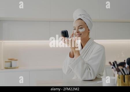 Donna felice in accappatoio bianco con asciugamano sulla testa cura della sua pelle dopo la doccia del mattino Foto Stock