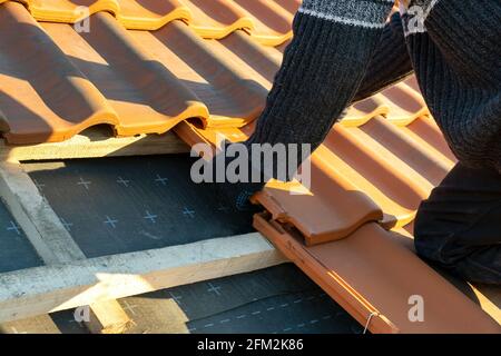 Closeup di mani di lavoro che installano tegole di ceramica gialla montate su tavole di legno che coprono il tetto dell'edificio residenziale in costruzione. Foto Stock