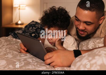 Il giovane padre e il figlio si stendano su un letto di sorveglianza un film su un tablet digitale alla sera Foto Stock