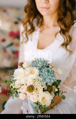Una sposa in un abito di pizzo si siede su un divano in una camera d'albergo e tiene un bouquet di nozze nelle sue mani, da vicino Foto Stock