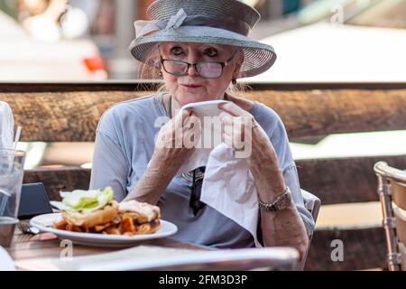 05-02-2021 Annapolis, MD, USA: Una vecchia signora caucasica che indossa un ampio cappello da sposa sta pranzando da sola in un ristorante. Lei sta strofinando la bocca Foto Stock