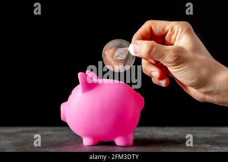 Immagine concettuale per l'utilizzo della valuta crittografica digitale per il risparmio. Una donna caucasica sta mettendo una moneta simbolica di Ethereum (ETH) in una banca di piggy come un risparmio Foto Stock
