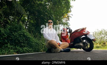 Uomo in moto rossa in abiti bianchi guida su strada forestale viaggio. Un uomo turista caucasico in occhiali da sole sedersi sul marciapiede, relax, vicino scooter. Foto Stock