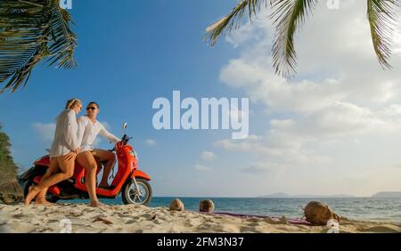 Viaggio su strada in scooter. Bella coppia su moto rossa in abiti bianchi sulla spiaggia di sabbia. Persone che camminano vicino alle palme tropicali, mare. Noleggio moto. Foto Stock