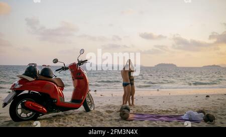 Viaggio su strada in scooter. Bella coppia su moto rossa in abiti bianchi sulla spiaggia di sabbia. Persone che camminano vicino alle palme tropicali, mare. Noleggio moto. Foto Stock