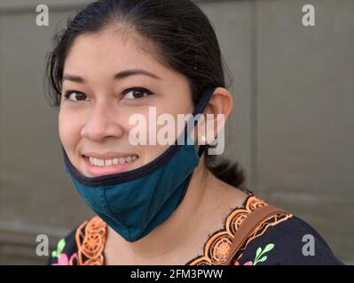 Affascinante giovane donna messicana dagli occhi marroni tira giù la sua maschera di tessuto non medico blu durante la pandemia globale del coronavirus e sorride alla macchina fotografica. Foto Stock