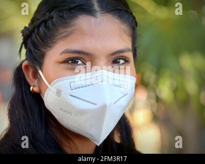 Giovane donna latina messicana positiva con bei occhi marroni indossa una maschera facciale KN95 durante la pandemia del coronavirus globale e guarda lo spettatore. Foto Stock