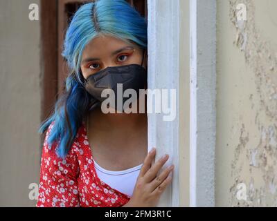 La ragazza teen di Latina caucasica con i capelli lunghi tinti blu e il make-up elaborato dell'occhio indossa una maschera nera del viso e porge intorno all'angolo di una porta d'ingresso. Foto Stock