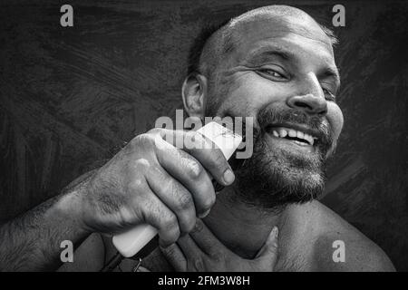 Uomo caucasico che cerca di radersi con un rasoio elettrico. Un brutale uomo calvo tiene un rasoio in mano e rade i peli corti su uno sfondo metallico Foto Stock