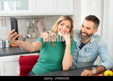 La coppia felice prende un selfie nella cucina. Bionda donna scatta una foto con il suo ragazzo. Smartphone, felicità e stile di vita domestico. Foto Stock