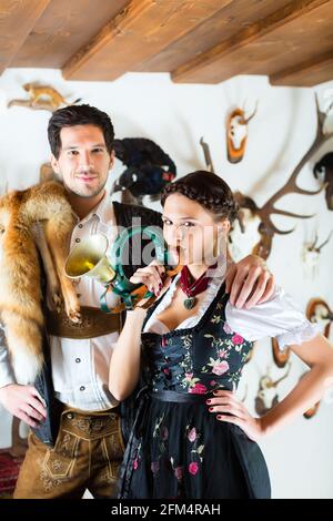 Giovane cacciatore con sua moglie e una tromba davanti a una parete con diversi corna, palchi e trofei in una baita alpina Foto Stock