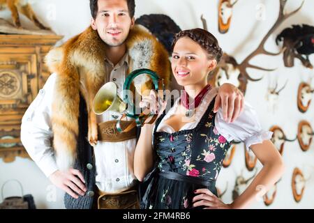 Giovane cacciatore con sua moglie e una tromba davanti a una parete con diversi corna, palchi e trofei in una baita alpina Foto Stock