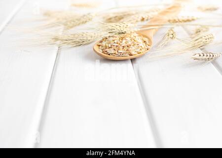 Fiocchi di grano in cucchiaio di legno con spighe di grano su tavola di legno bianco. Cereale intero con crusca di grano. Spazio di copia Foto Stock