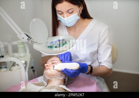 Medico cosmetologo in guanti di gomma sterile e maschera, utilizzando scrubber ultrasonico durante la pulizia del viso della giovane ragazza. Pulizia professionale, trattamento della pelle problematico Foto Stock