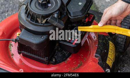 Primo piano di un uomo caucasico che riempie il serbatoio del gas di un tosaerba rosso con benzina proveniente da un beccuccio giallo. Tagliaerba e erba sul rasaerba. Foto Stock