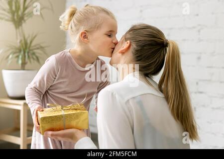 figlia piccola che dà il presente e il bacio alla sua mamma a casa. compleanno, giorno delle donne o concetto di giorno delle madri Foto Stock