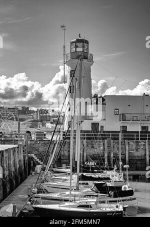 Gli yacht in una linea sono in primo piano che conducono ad un faro bianco. Edifici e una ruota di ferro sono sullo sfondo. Foto Stock