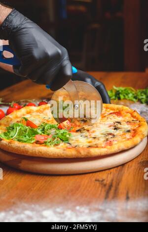 Primo piano verticale foto di uno chef in guanti di gomma nera affettando la pizza sul tagliere con farina sparsa intorno al piatto. Foto Stock