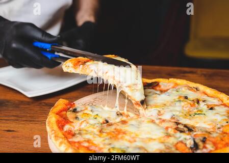 Vista ravvicinata dello chef in guanti di gomma nera che raccoglie una fetta di pizza da forno a caldo e sciogliente dal piatto di legno. Foto Stock