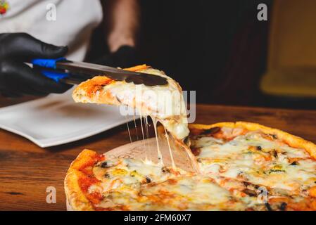 Vista ravvicinata dello chef in guanti di gomma nera che raccoglie una fetta di pizza da forno a caldo e sciogliente dal piatto di legno. Foto Stock