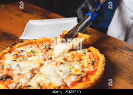 Vista ravvicinata dello chef in guanti di gomma nera che raccoglie una fetta di pizza da forno a caldo e sciogliente dal piatto di legno. Foto Stock