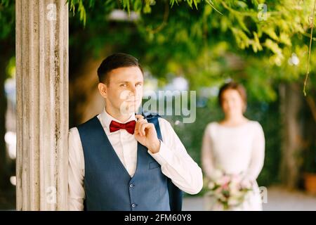 Lo sposo sorridente gettò la giacca sopra la spalla. Lo sposo si appoggiò contro una colonna nel giardino Foto Stock