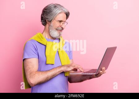 Profilo Vista laterale ritratto di un'attraente presa uomo con capelli grigi focalizzata utilizzo di un computer portatile per la scrittura di messaggi di posta elettronica isolati su sfondo rosa color pastello Foto Stock