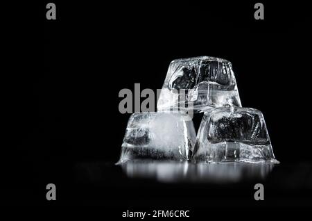 Tre cubetti di ghiaccio parzialmente fusi impilati su un riflettente nero Superficie e sfondo nero Foto Stock