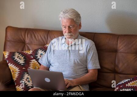 Un uomo grigio con capelli grigi, anziano e insoddisfatto, si siede sul divano guardando lo schermo del dispositivo lavorando su un computer portatile seduto da solo a casa. Vecchio uomo d'affari che scrive pasticcio Foto Stock