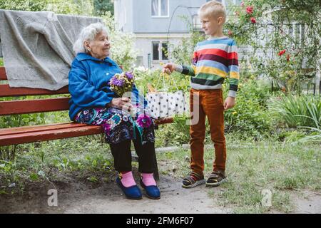 bisnonna di 90 anni, nonna con nipote insieme. Nipote abbraccia la sua amata nonna. Il nipote di capretto è venuto alla nonna con il fiore Foto Stock