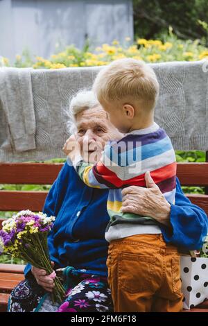 bisnonna di 90 anni, nonna con nipote insieme. Nipote abbraccia la sua amata nonna. Il nipote di capretto è venuto alla nonna con il fiore Foto Stock