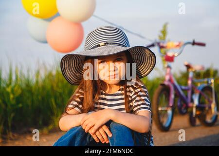 Carino ragazza poco infelice in cappello grande del sole che siede sopra strada con la sua bicicletta dietro di lei Foto Stock