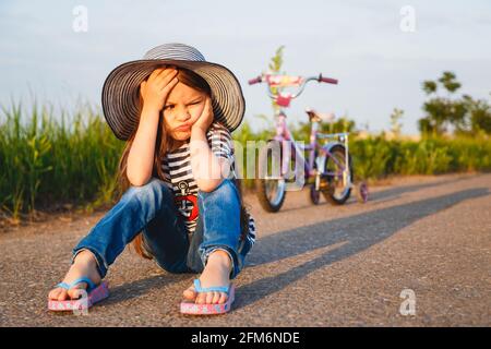 Carino ragazza poco infelice in cappello grande del sole che siede sopra strada con la sua bicicletta dietro di lei Foto Stock